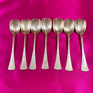 May include: Set of eight silver spoons with a rounded bowl and a flared handle. The spoons are arranged in a row on a pink satin surface.