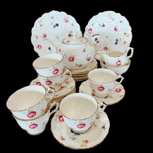 May include: A vintage tea set comprising a teapot, cups, saucers, plates, and a creamer. The white porcelain is adorned with a pattern of red apples, purple berries, and green leaves, with gold edging. A classic tea service.