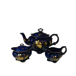 Pode incluir: Um conjunto de bule, jarra de creme e açucareiro em azul cobalto com detalhes florais dourados. O conjunto é feito de cerâmica e tem um estilo vintage.