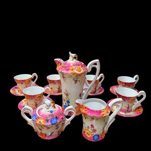 Puede incluir: Un juego de té vintage con un diseño floral. El juego incluye una tetera, una jarra de crema, un azucarero y varias tazas y platillos. El juego es principalmente blanco con detalles florales rosas, naranjas y azules. Las asas y los bordes son dorados.