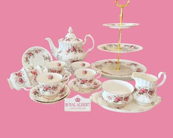 Peut inclure: Un service à thé Royal Albert en porcelaine blanche orné de motifs de roses roses et de bordures dorées. L'ensemble comprend une théière, un présentoir à gâteaux à trois étages, plusieurs tasses avec soucoupes, un sucrier et un pot à lait.
