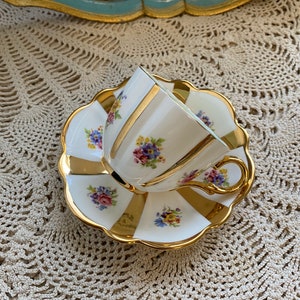 May include: A white teacup and saucer with a gold rim and floral pattern. The saucer has a scalloped edge.