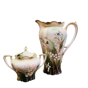 May include: A vintage porcelain pitcher and sugar bowl set with a delicate floral design. The white porcelain is decorated with green leaves and white lily of the valley flowers.