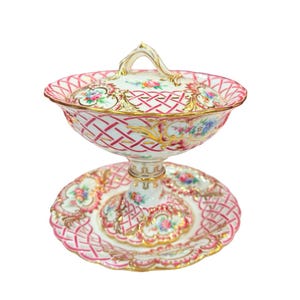 May include: A decorative porcelain serving dish with a lid, featuring a pink lattice pattern and floral accents. The dish sits on a matching pedestal base with a similar design, and gold trim. The handle on the lid is gold.