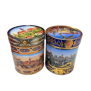May include: Two cylindrical ceramic containers with lids, each decorated with detailed landscape paintings of European towns and castles. The lids and rims are trimmed with gold and blue patterns. One container has a red and gold design, the other a blue and gold design.