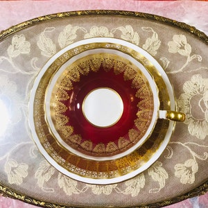 May include: A red and gold teacup and saucer with a delicate lace pattern. The cup and saucer are on a white lace doily.