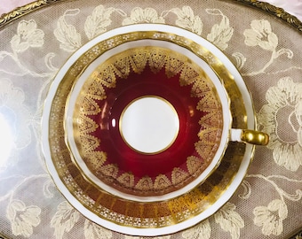 May include: A red and gold teacup and saucer with a delicate lace pattern. The cup and saucer are on a white lace doily.