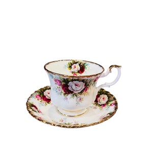 Op de afbeelding: Een wit theekopje en schotel met een bloemenpatroon met roze, rode en witte rozen. Het kopje en de schotel hebben een gouden rand.