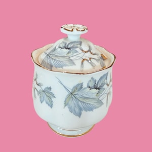 May include: A white porcelain sugar bowl with a lid, decorated with a leaf pattern in grey and blue, and gold trim. The bowl has a rounded shape and a flower-shaped handle. The background is a solid pink colour.