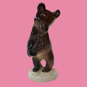 Puede incluir: Una figura de cerámica brillante de un oso pardo de pie sobre sus patas traseras. El oso tiene un pelaje marrón oscuro con tonos más claros en el pecho y la cara, y está colocado sobre una base circular de color blanco roto con acabado liso.