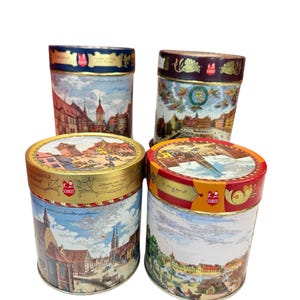 May include: Four vintage Schmidt biscuit tins with illustrations of European city scenes. The tins are cylindrical with gold and red detailing. Each tin features a different historical townscape, with text on the lids.