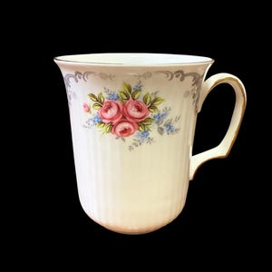 May include: Cream-colored ceramic mug with a floral design featuring pink roses and blue accents. The mug has a gold-trimmed handle and rim, and a vertical ribbed texture. The mug is set against a black background.
