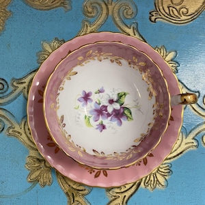 May include: A pink teacup and saucer with a gold trim and floral design. The cup and saucer have a scalloped edge. The floral design features purple and white flowers.