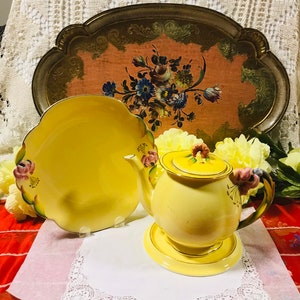 Puede incluir: Una tetera amarilla con detalles florales y un plato a juego en una bandeja con un diseño floral. La tetera tiene una tapa y un asa. El plato es redondo y tiene un borde festoneado.