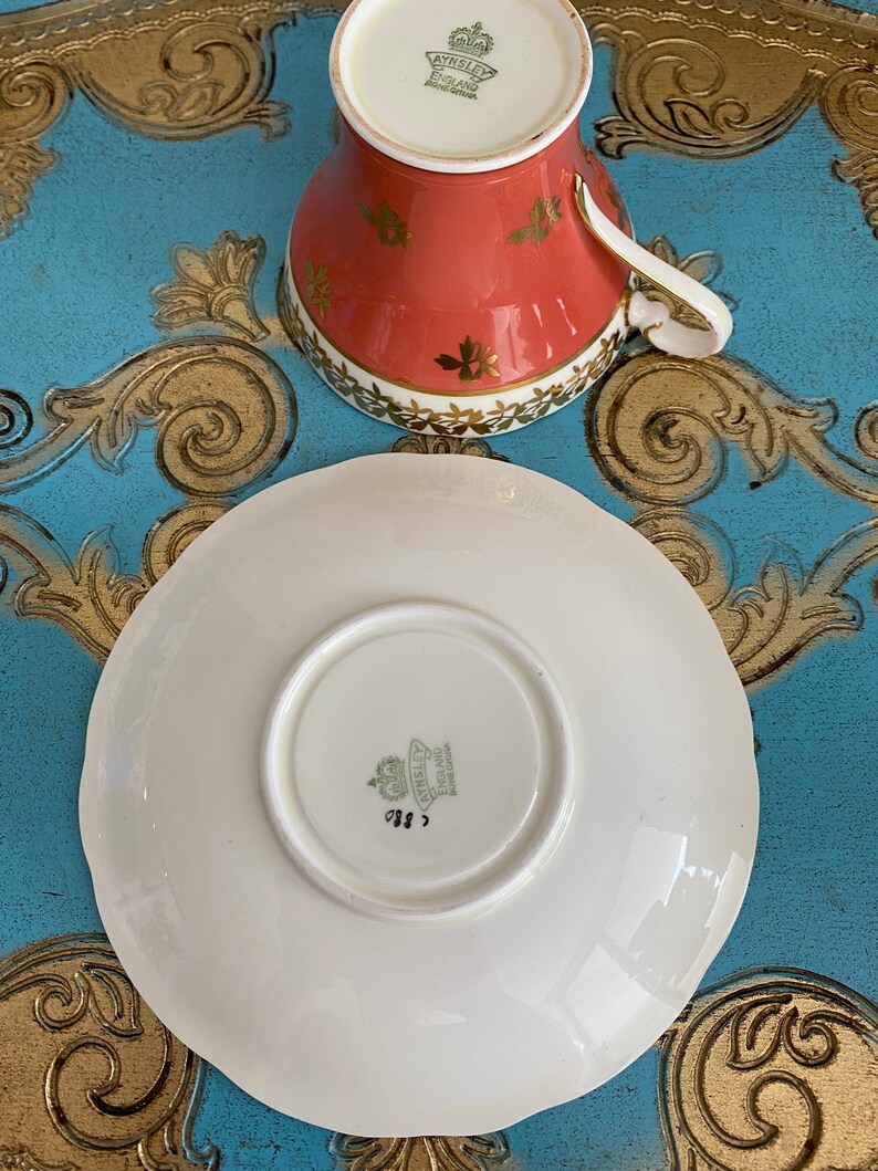 May include: A white teacup and saucer with a gold rim and floral design. The saucer has a scalloped edge. The teacup and saucer are marked with the Aynsley England Bone China logo.