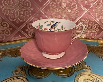 May include: A pink teacup and saucer with a gold trim. The teacup features a floral design with a blue bird.