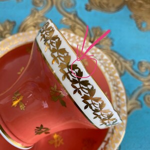 May include: A coral-colored teacup and saucer with a white rim and gold floral pattern. The cup and saucer are decorated with a delicate gold floral design.