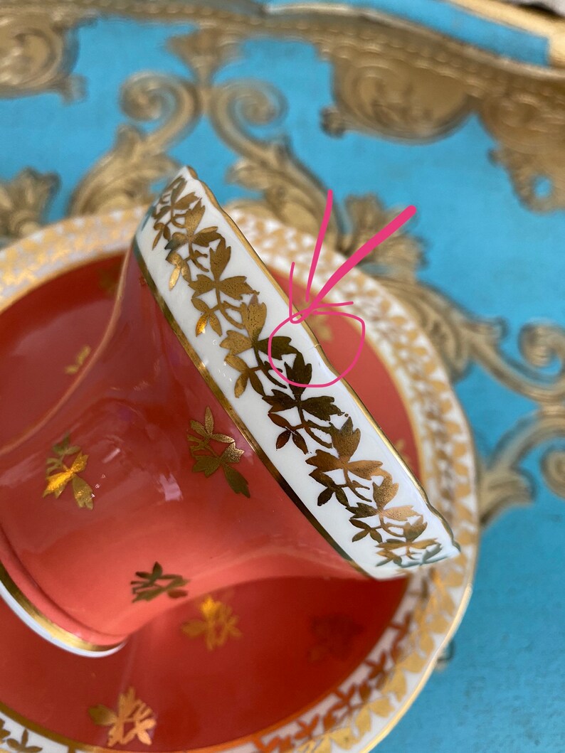 May include: A coral-colored teacup and saucer with a white rim and gold floral pattern. The cup and saucer are decorated with a delicate gold floral design.