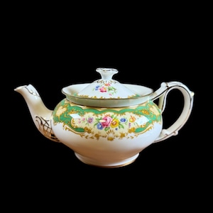 Peut inclure: Une théière en porcelaine de couleur crème avec un couvercle. La théière présente un motif floral vert et or autour du milieu et sur le couvercle. La poignée et le bec sont de couleur crème avec des détails dorés. La théière est sur fond noir.