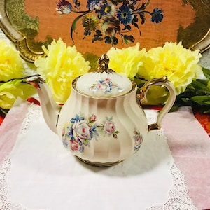 Puede incluir: Una tetera blanca con un diseño floral y detalles dorados. La tetera tiene una tapa con un pomo y un pico. La tetera está sobre un encaje blanco.