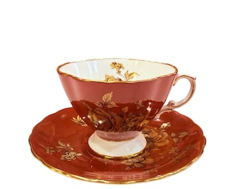 Può includere: Un set vintage di tazza e piattino da tè. La tazza e il piattino sono di colore rosso scuro con disegni floreali dorati. La tazza ha un interno bianco e una base bianca. I bordi della tazza e del piattino sono rifiniti in oro.