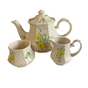 May include: A white teapot and two creamers with a floral pattern in shades of yellow, pink, blue, and green. The teapot and creamers have a woven pattern.