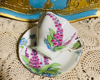 May include: A white teacup and saucer with a floral pattern. The cup and saucer feature pink and green flowers with blue accents. The cup and saucer are sitting on a white crocheted doily.