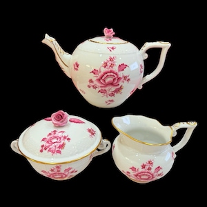 Puede incluir: Un juego de té de porcelana de tres piezas con tetera, azucarero con tapa y jarra de crema. El juego es blanco con diseños florales rosas y ribetes dorados. La tetera tiene un adorno en forma de rosa en la tapa.