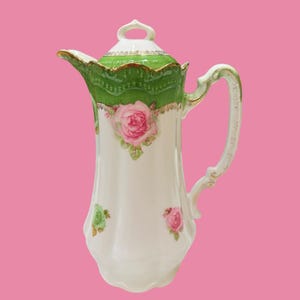 Op de afbeelding: Een witte porseleinen koffiepot met een geschulpte groene rand met gouden afwerking en een bijpassend deksel. Het lichaam is versierd met handgeschilderde roze rozen en groene bladeren, aangevuld met een gebogen handvat en een tuit.
