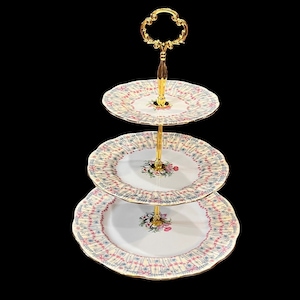 Może przedstawiać: Trójpoziomowa podstawka na ciasto z pozłacanym metalowym stelażem i białą porcelanową podstawą. Każdy poziom zdobi delikatny wzór kwiatowy w odcieniach różowego, niebieskiego i żółtego.