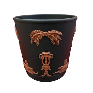 May include: Black planter with embossed wheat and lyre designs in a terracotta color. The planter has a wide opening and a slightly tapered shape. The decorative elements are symmetrically arranged around the planter.