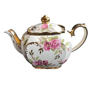 Può includere: Una teiera bianca con un bordo dorato e un motivo floreale di rose rosa. La teiera ha un coperchio con un pomo dorato.