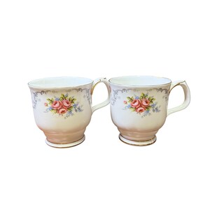 Op de afbeelding: Twee witte keramische mokken met een roze basis en gouden rand. Elke mok heeft een bloemenontwerp met roze rozen en blauwe accenten. De mokken hebben gebogen handvatten.