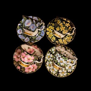May include: Set of four teacups and saucers with floral designs. Each cup and saucer features a different floral pattern: blue morning glories, yellow flowers, pink flowers, and white flowers. The cups and saucers have a black background with gold accents.