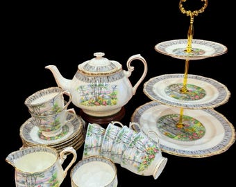 May include: A complete vintage tea set, including a teapot, cups, saucers, a creamer, a sugar bowl, and a three-tiered cake stand. The set has a landscape design with a white fence, trees, and flowers, with gold trim on a light blue border.