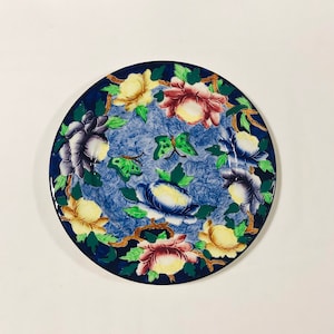 Puede incluir: Un plato de cerámica azul y blanco con un diseño floral. El plato presenta un borde de flores amarillas, rosas y moradas con hojas verdes. Hay dos mariposas verdes en el centro del plato.