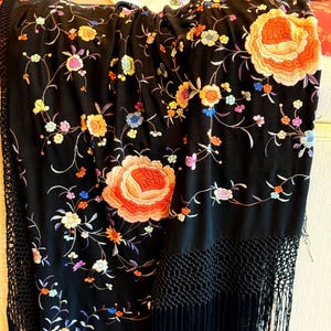 May include: A black silk shawl with intricate floral embroidery in shades of orange, pink, yellow, blue, and green. The shawl has a black fringe and a delicate net pattern along the bottom edge.