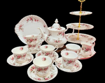 May include: A classic tea set with white porcelain items decorated with pink rose patterns. The set includes a teapot, cups, saucers, a creamer, a sugar bowl, and a three-tiered serving stand. The edges of the plates and saucers have a scalloped design.