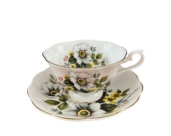 May include: A delicate teacup and saucer set. The white porcelain cup and saucer feature a floral design with white and yellow flowers, green leaves, and gold trim. The cup has a curved handle and a scalloped rim.