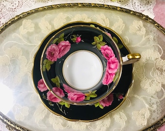 Könnte beinhalten: Eine schwarze Teetasse und Untertasse mit goldfarbenem Rand und einem Blumendesign. Die Blumen sind rosa und weiße Rosen mit grünen Blättern auf schwarzem Hintergrund.