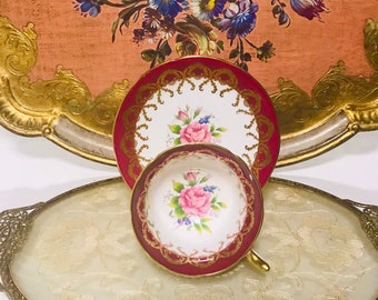 May include: A vintage teacup and saucer set with a floral design. The teacup and saucer are both white with a gold trim and a red rim. The teacup has a pink rose and green leaves design. The saucer has a floral design with a gold trim.