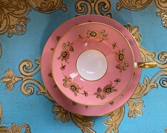 May include: A pink teacup and saucer with a gold floral pattern on a blue and gold tray.