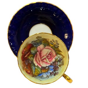 Peut inclure: Un service à thé vintage composé d'une tasse et d'une soucoupe. La tasse présente un motif floral avec une grande rose rose et d'autres fleurs colorées. La soucoupe est bleu foncé avec un bord doré.