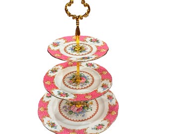 Puede incluir: Un soporte de tres niveles con platos de porcelana blanca que presentan bordes festoneados de color rosa y motivos florales. Los platos están unidos por una varilla central de metal dorado con un asa ornamentada en la parte superior.