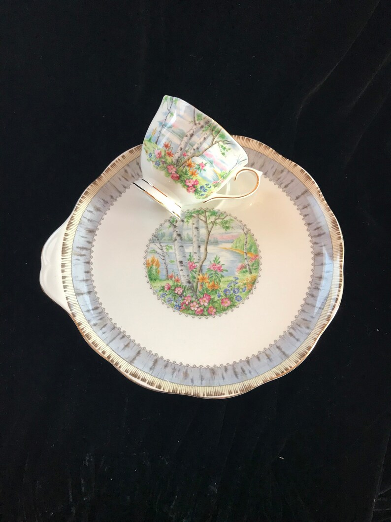 Pu&ograve; includere: Un set vintage di tazza e piattino da t&egrave;. La tazza e il piattino sono bianchi con bordi azzurri e dorati. La tazza e il piattino presentano un design paesaggistico con alberi, fiori e uno specchio d'acqua. La tazza &egrave; appoggiata sul piattino.