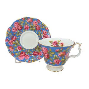 May include: A vintage teacup and saucer set. The blue china is decorated with pink and red roses. The teacup has a gold handle and base, and the saucer has a scalloped edge. A classic tea set.