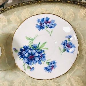 May include: A white teacup saucer with a gold rim and a floral design featuring blue and purple cornflowers.