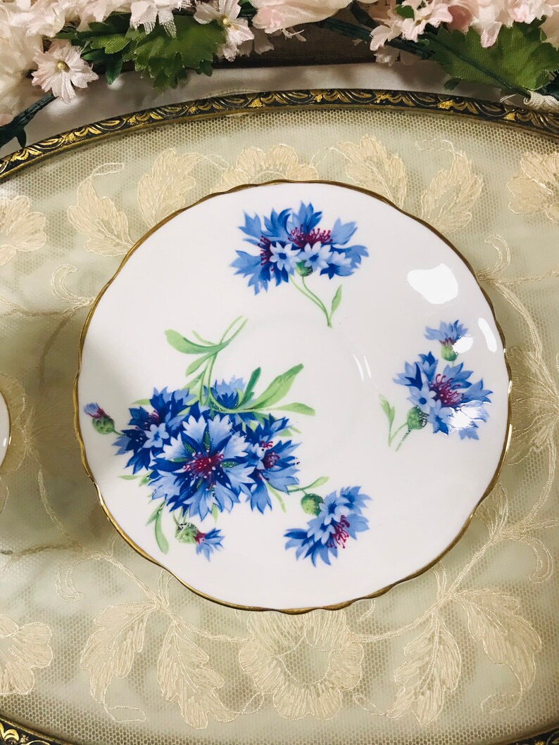 May include: A white teacup saucer with a gold rim and a floral design featuring blue and purple cornflowers.