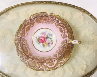 May include: A pink teacup and saucer with a gold floral design. The teacup has a floral pattern in the center and a gold rim.