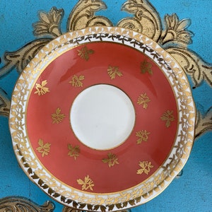 May include: A coral-colored saucer with a gold rim and a delicate floral pattern. The saucer is made of fine china and has a white center.